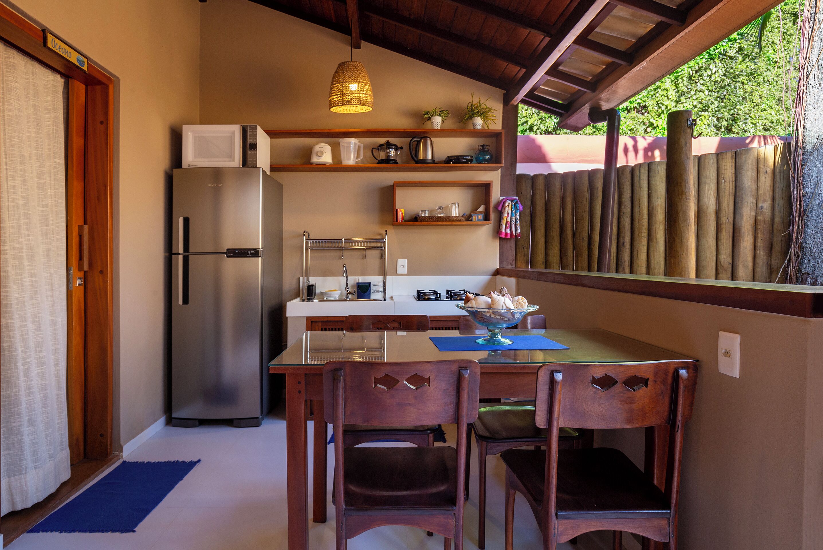 Cocina compacta con refrigerador, microondas y varios electrodomésticos en estantes. Hay sillas de madera y una mesa con un mantel individual.