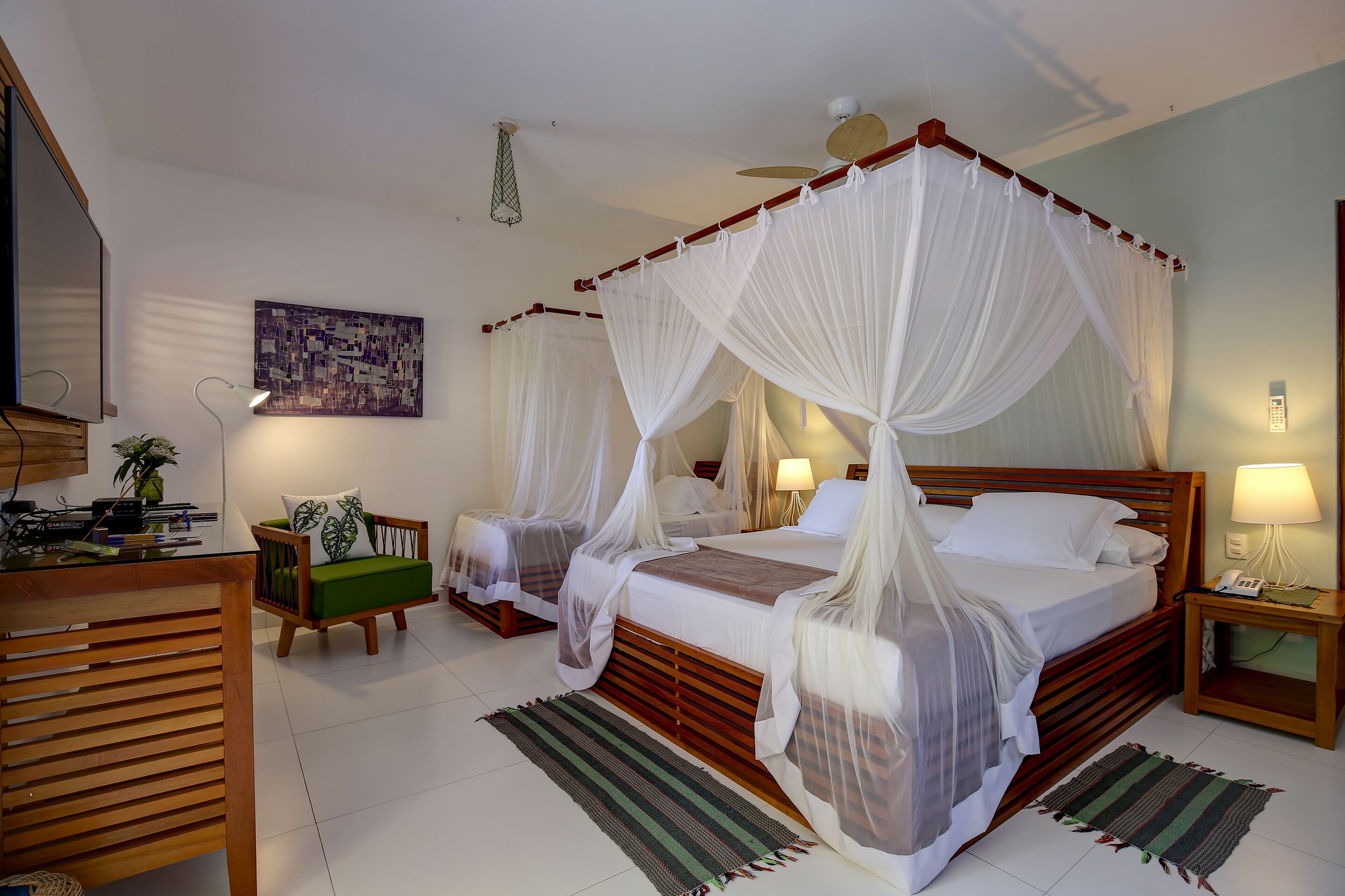 Una cama con dosel adornada con cortinas blancas, una mesita de noche de madera con una lámpara, una silla verde y un escritorio con una computadora y una planta.