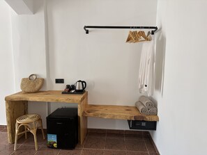 Une petite pièce comprenant un bureau en bois, un petit tabouret, une valise noire, une machine à café et un porte-serviettes avec des vêtements.