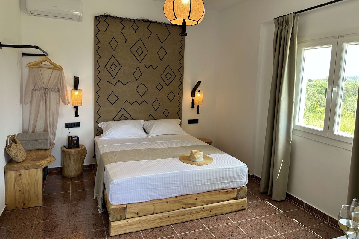 A bedroom with a bed, a hanging light fixture, a wall-mounted lamp, a window with curtains, and a wooden nightstand.