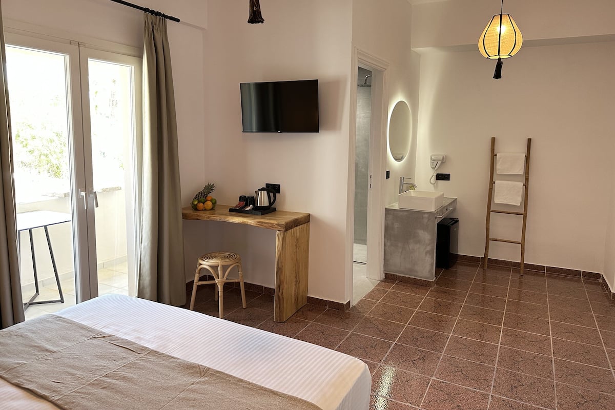 A hotel room with a bed, a desk with a computer and a chair, a television mounted on the wall, a bathroom with a mirror and a towel rack, and a wooden ladder.