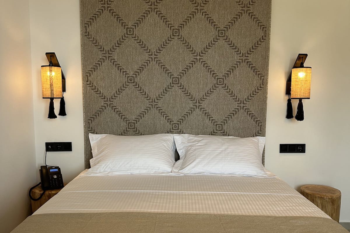 A neatly made bed with a patterned headboard, two wall-mounted lamps, a bedside phone, and a small wooden stool.