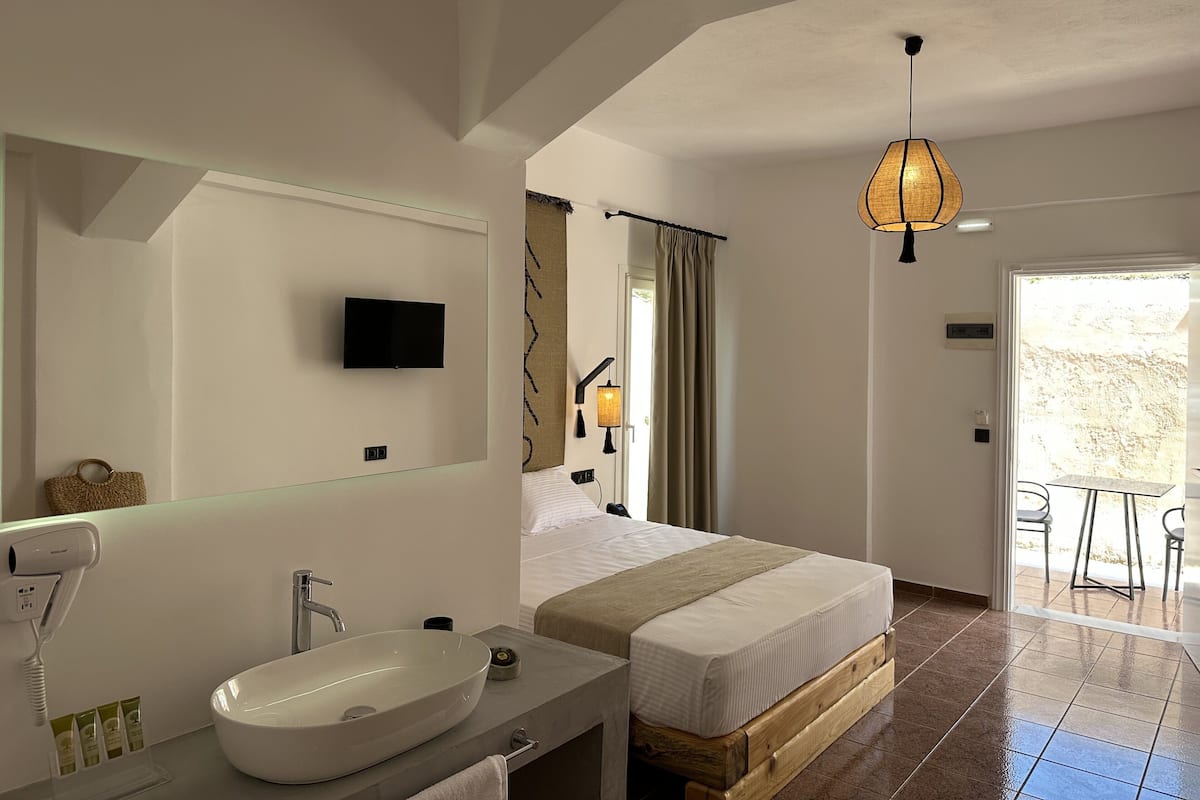 A modern hotel room with a large bed, a bathroom with a sink and mirror, a television, and a balcony with chairs.