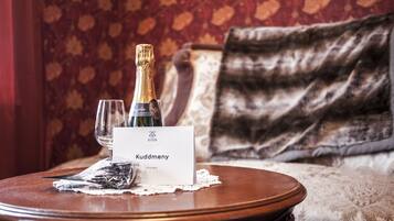 A bottle of champagne, a glass, and a name card on a round wooden table in a room with patterned wallpaper and a sofa.