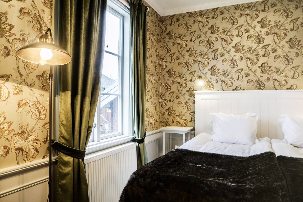 A room with a bed, a nightstand, a lamp, and a window with curtains.