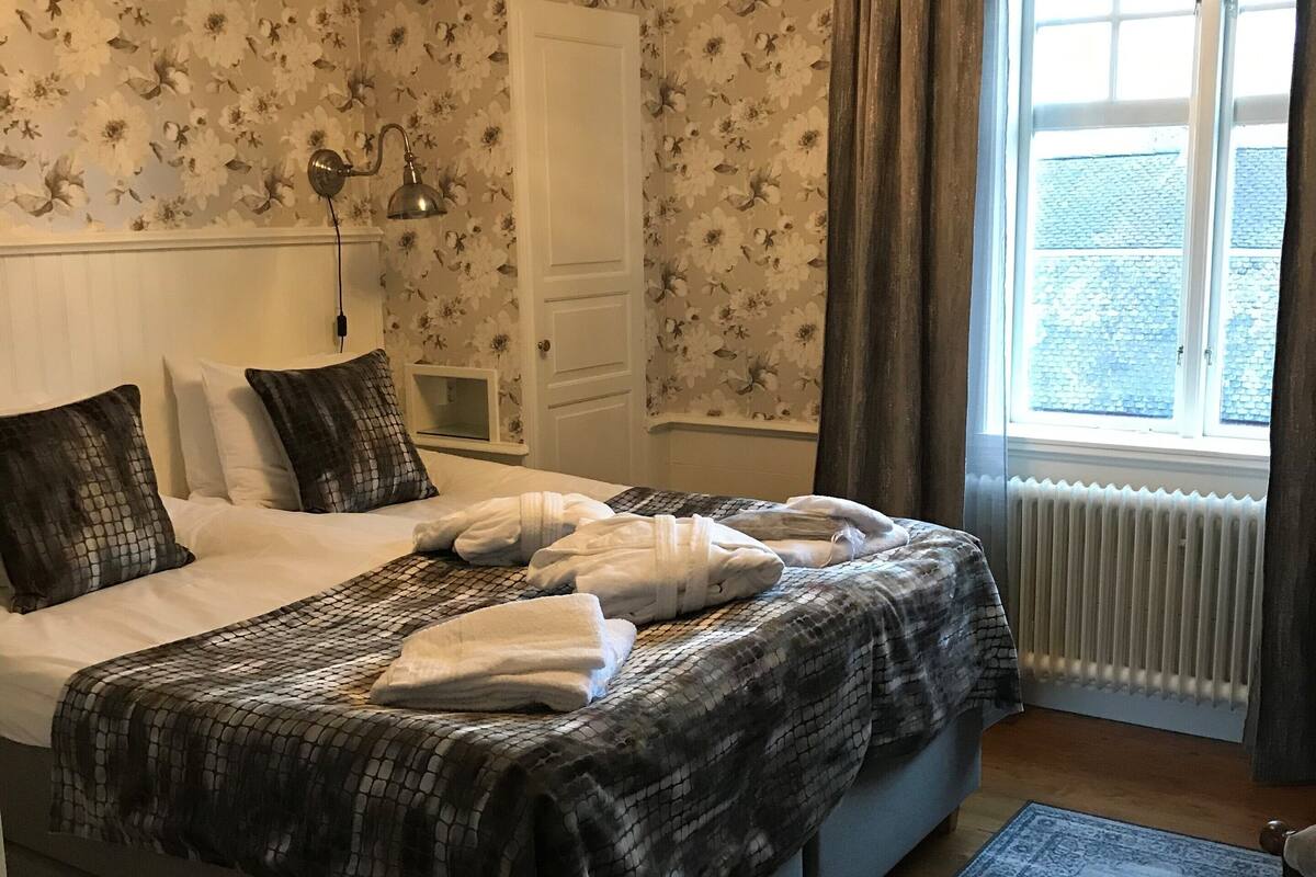 A bedroom with a bed, a nightstand, a lamp, a window with curtains, and a radiator.