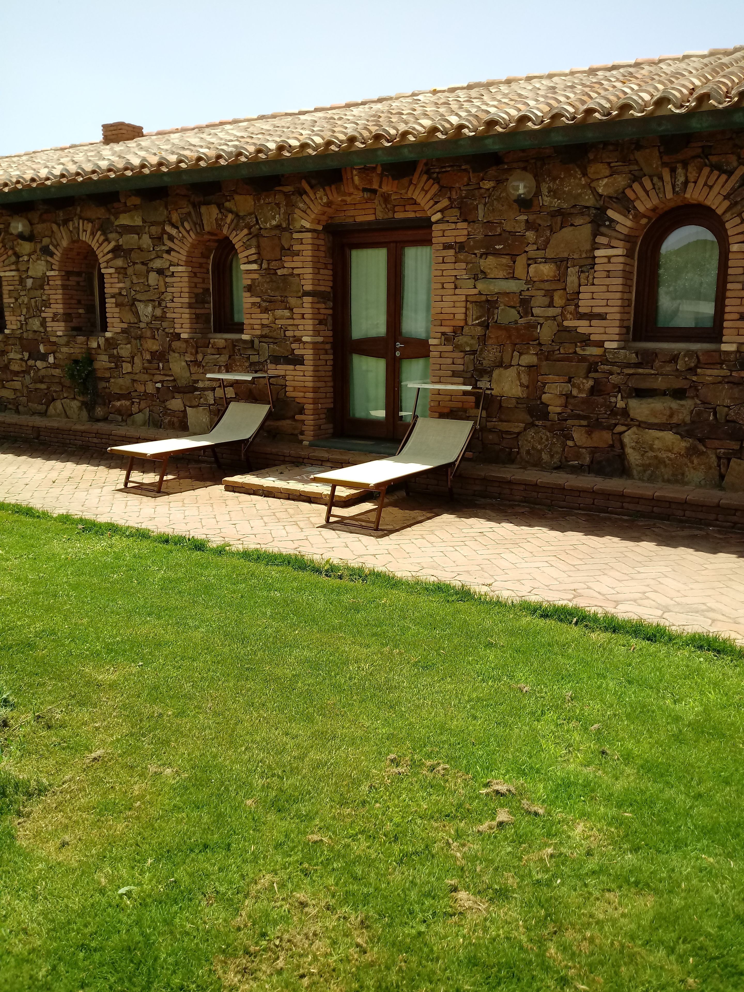 A stone building with a tiled roof, two lounge chairs, and a paved pathway leading to a grassy area.