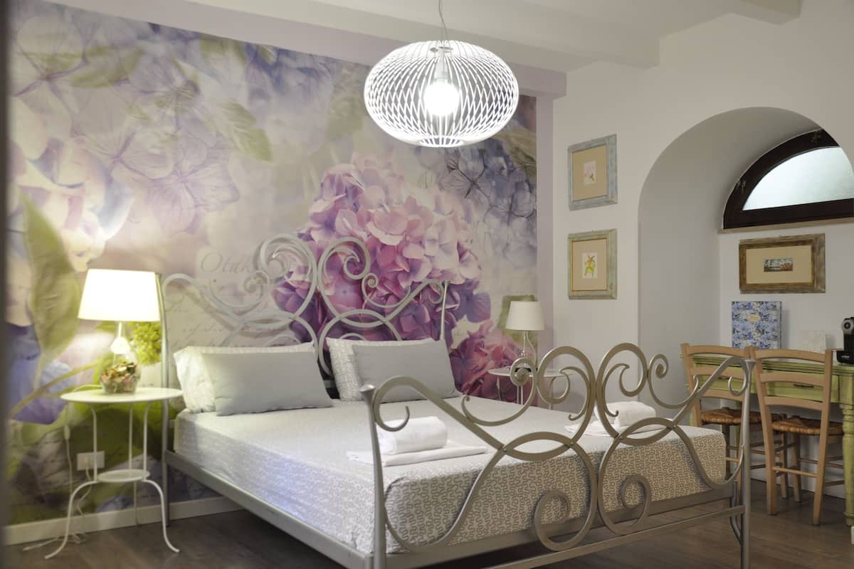 A bedroom with a bed, a chandelier, a wall with floral wallpaper, a small table with a lamp, and a dining area with chairs and a table.