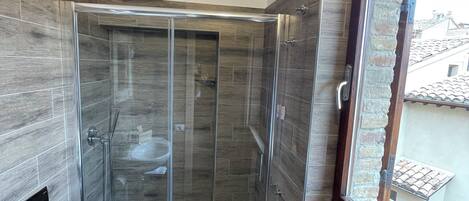 Un baño moderno con ducha envuelta en vidrio, inodoro y ventana con vistas a edificios.
