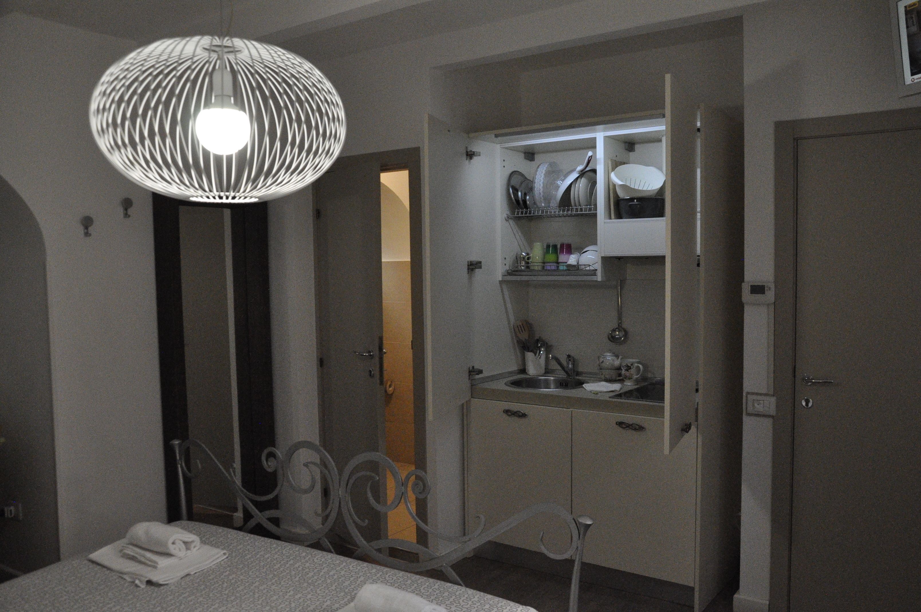 A bedroom with a bed, a bedside table, a hanging light fixture, a bathroom with a sink, and a cabinet with dishes.