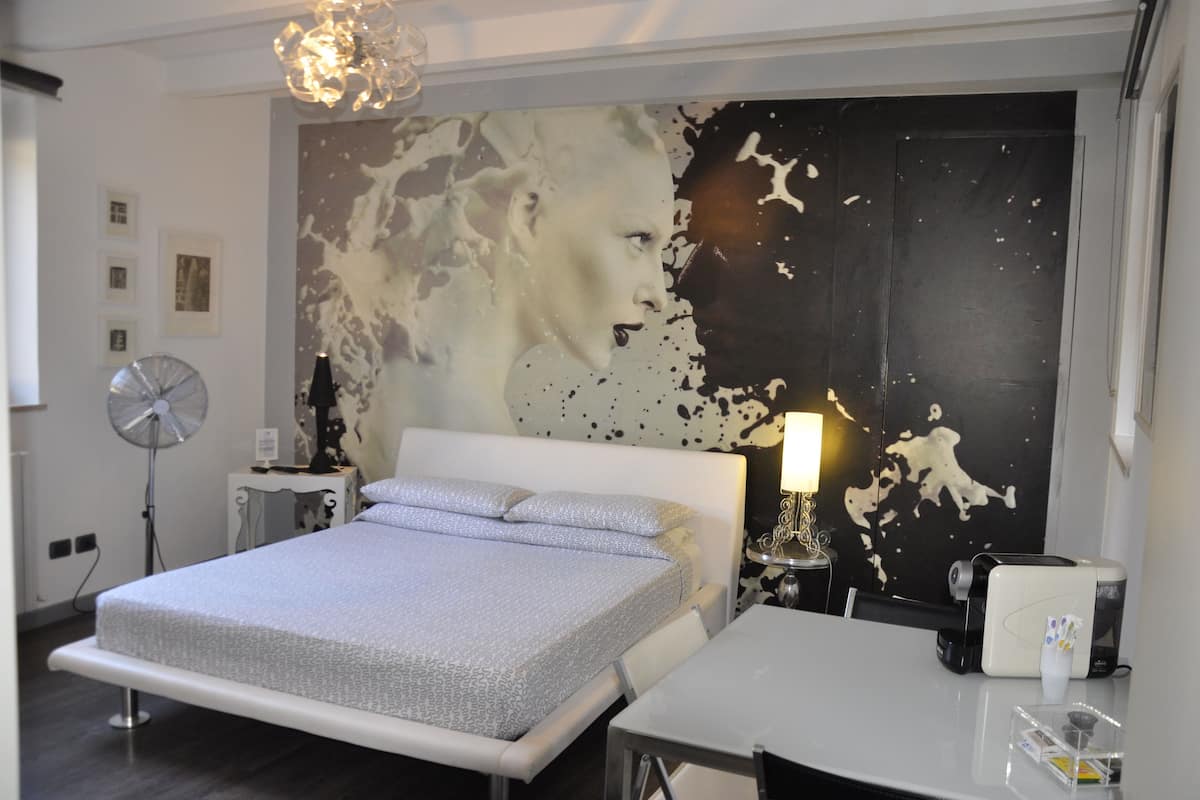 A modern bedroom with a large bed, a bedside table with a lamp, a wall-mounted fan, and a decorative wall piece.