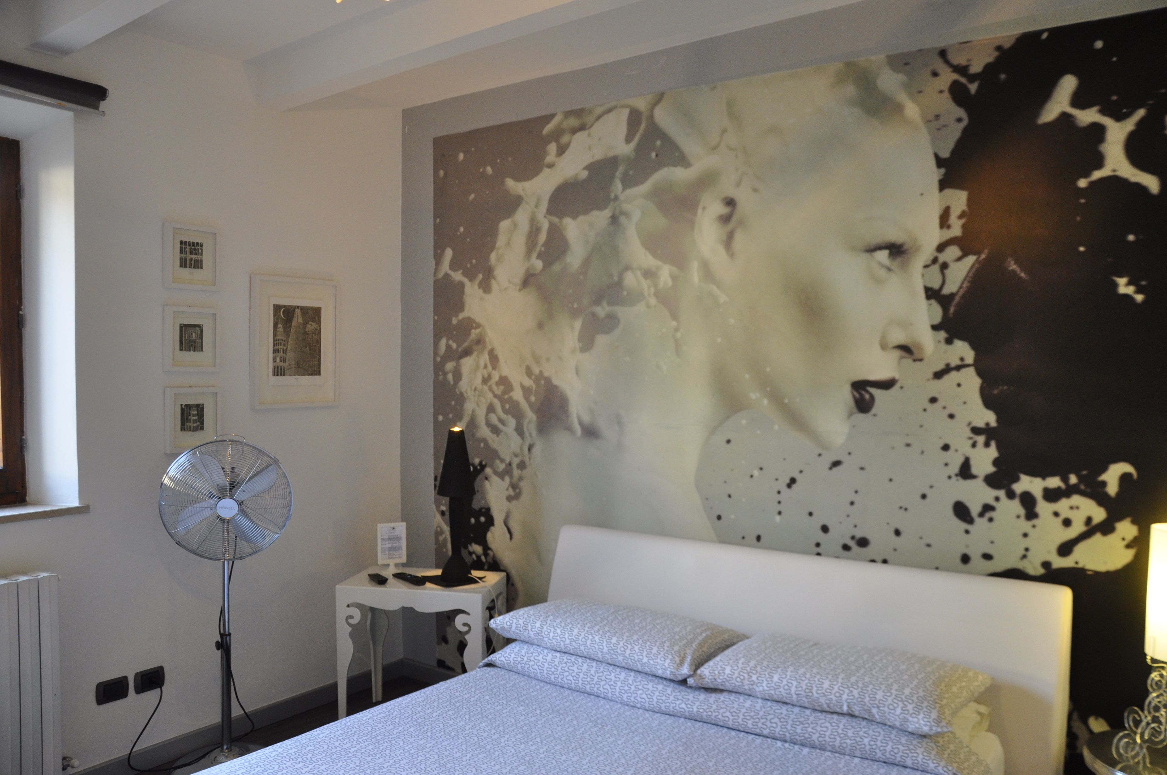 Une chambre avec un grand tableau abstrait accroché au mur, un ventilateur sur pied, une table de chevet avec une lampe et un lit soigneusement fait.