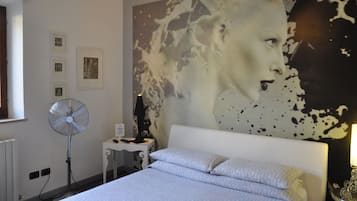 Un dormitorio con un cuadro abstracto grande en la pared, un ventilador de pie, una mesita de noche con lámpara y una cama bien hecha.