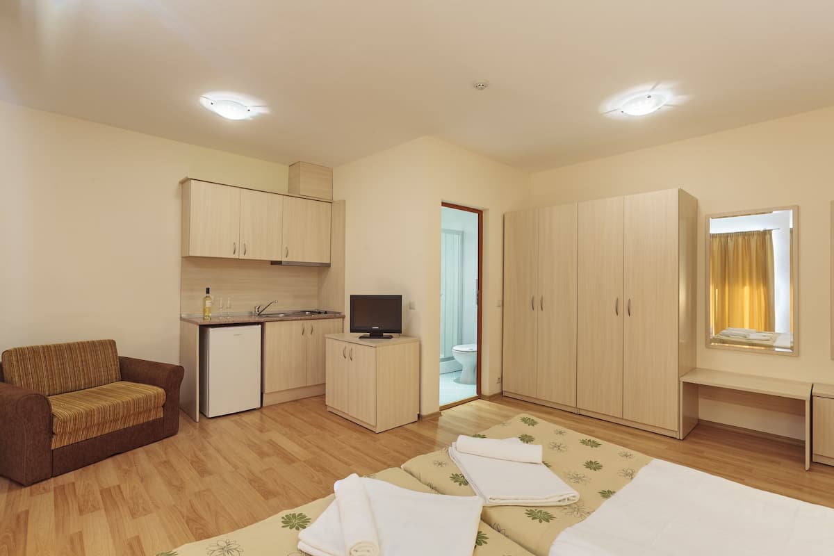 A compact hotel room with a kitchenette, a sofa, a bed with a floral bedspread, and a wardrobe.