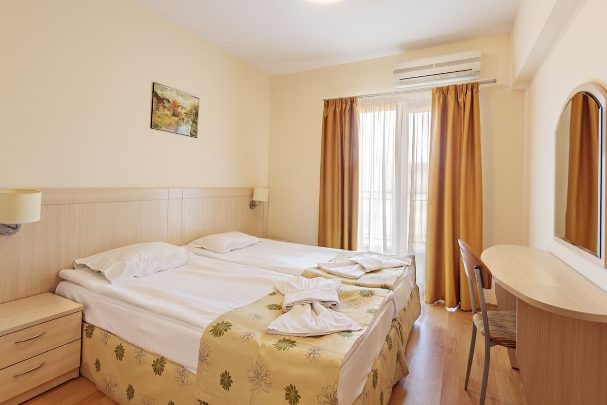 A hotel room with two beds, a desk, a chair, a lamp, and an air conditioning unit.