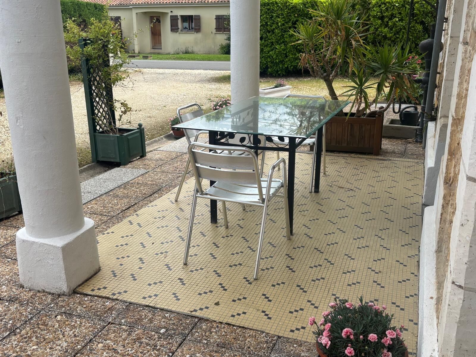 Une terrasse extérieure avec une table en verre et des chaises en métal, entourée d’un sol carrelé et de verdure.