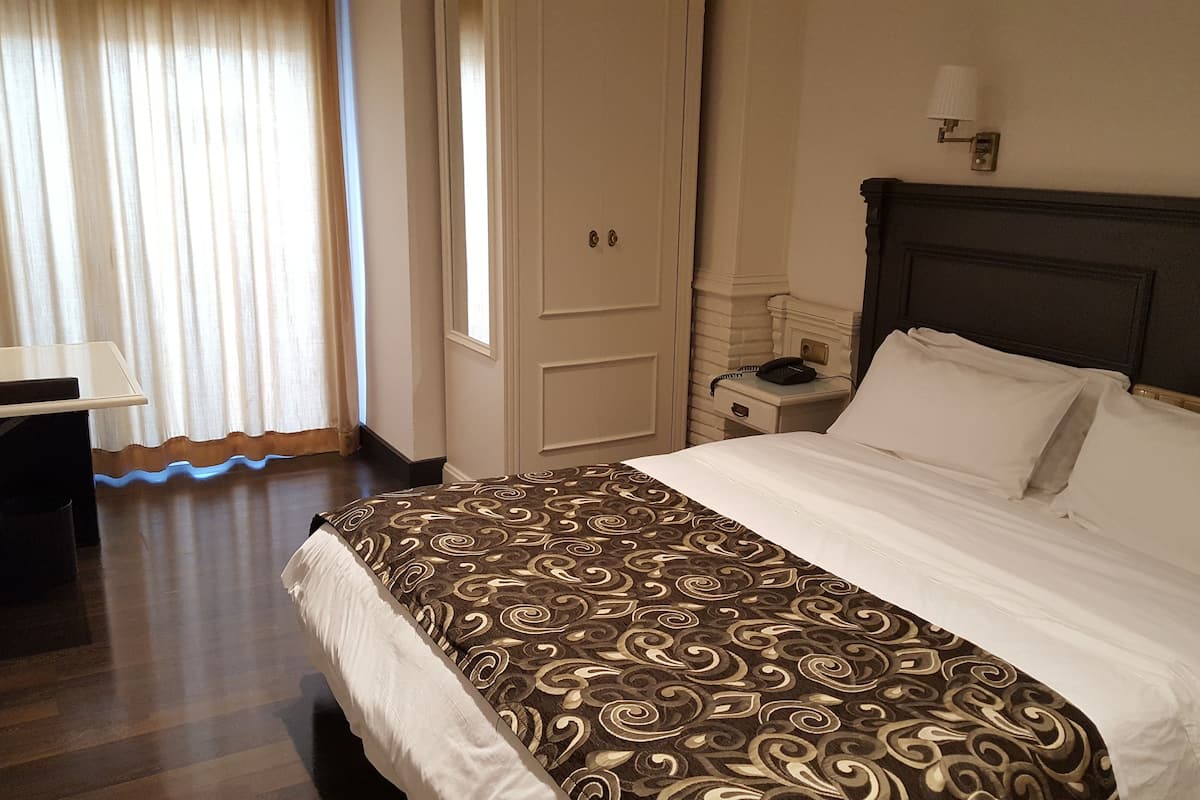 A hotel room with a bed, a nightstand, a lamp, a desk with a telephone, and a window with curtains.