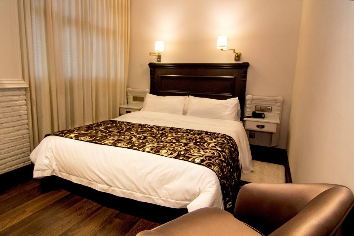 A hotel room with a bed, two bedside lamps, a nightstand, a chair, and a white wall with a brick-like pattern.