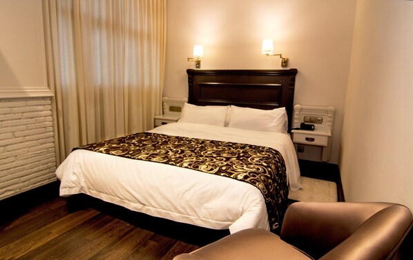 Habitación de hotel con cama, dos lámparas de noche, mesita, una silla y pared blanca con diseño tipo ladrillo.