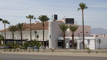 A modern hotel with a flat roof, palm trees, and a clear sky.