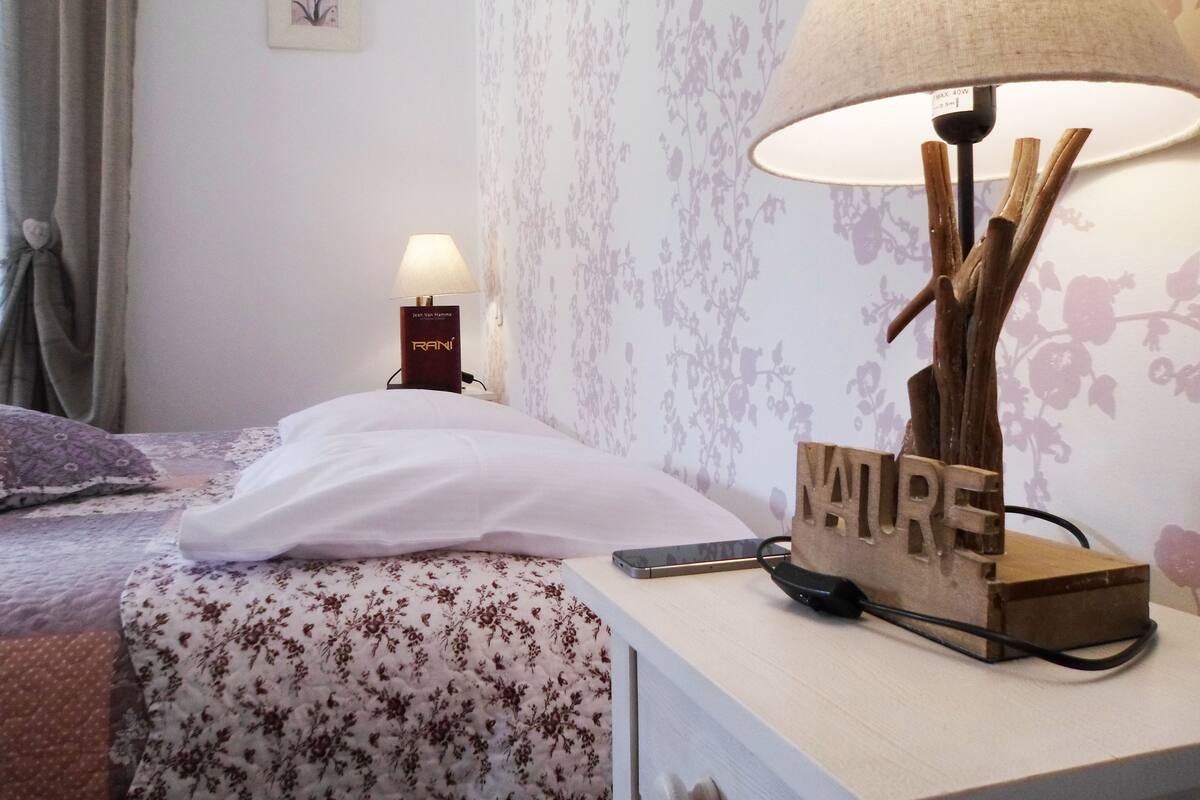 Una cama bien hecha con una colcha de estampado floral, una lámpara de noche con base de madera y un marco de pared.