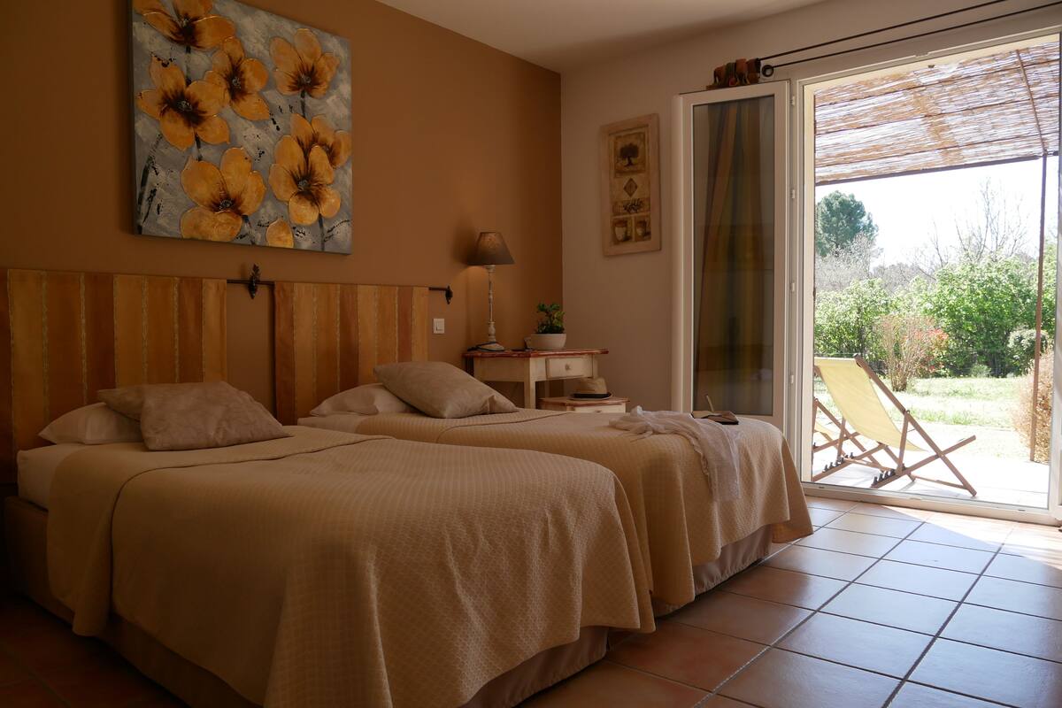Habitación de hotel con dos camas, un ventanal con vistas al exterior, un cuadro en la pared y una mesita con una lámpara y un sombrero.