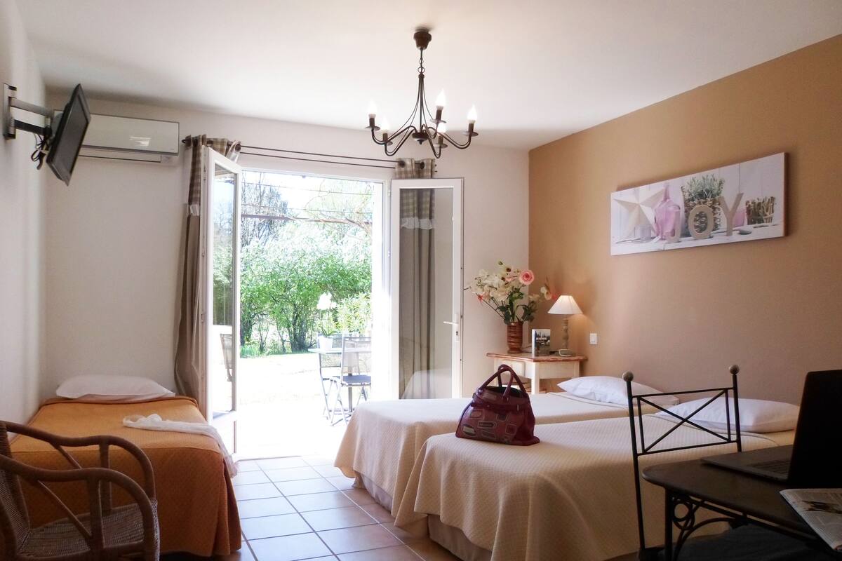 Habitación de hotel con dos camas, un escritorio con una computadora portátil, una silla, un televisor, una araña y una mesa con una lámpara, además de un ventanal con cortinas.