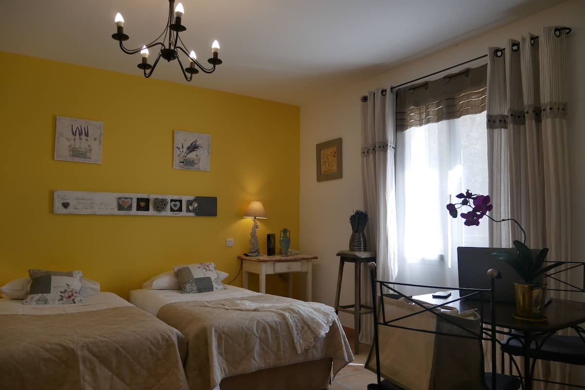 Habitación de hotel con dos camas, un candelabro, un escritorio con una silla y una ventana con cortinas.
