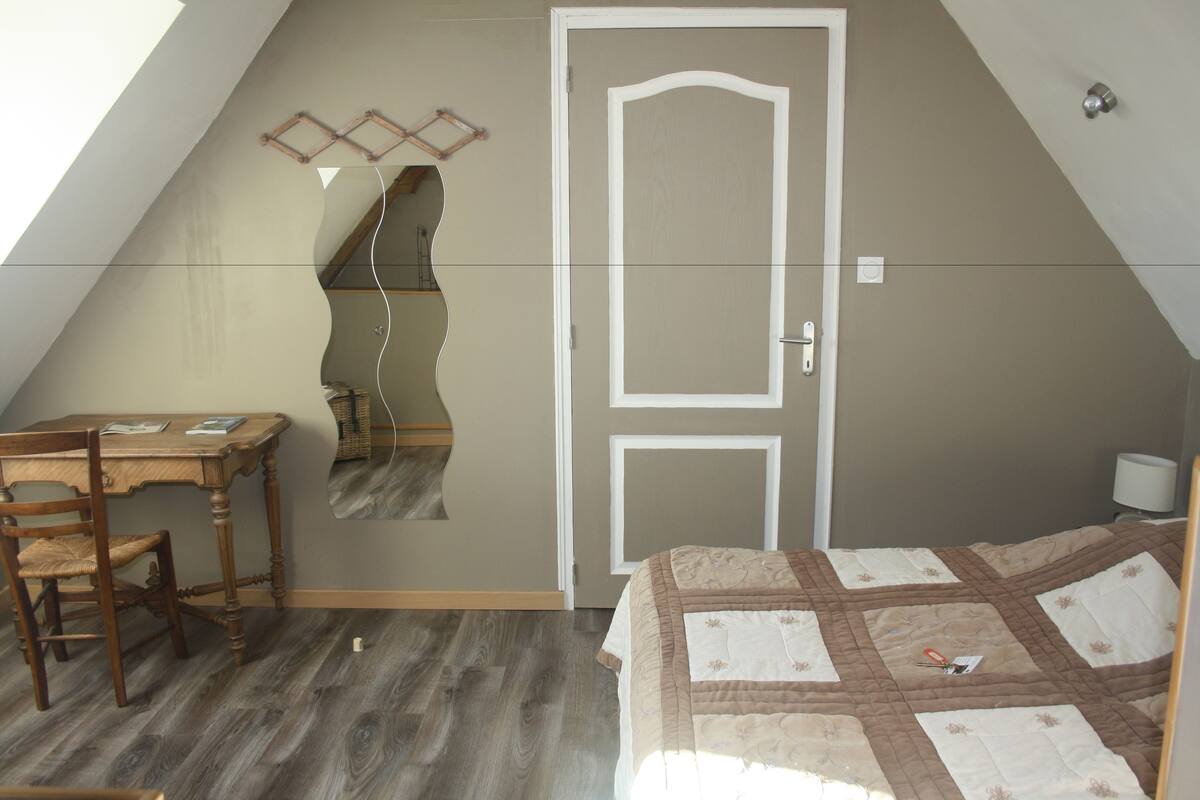 Un dormitorio con techo inclinado, un escritorio de madera, una silla, una cama con colcha y una puerta.