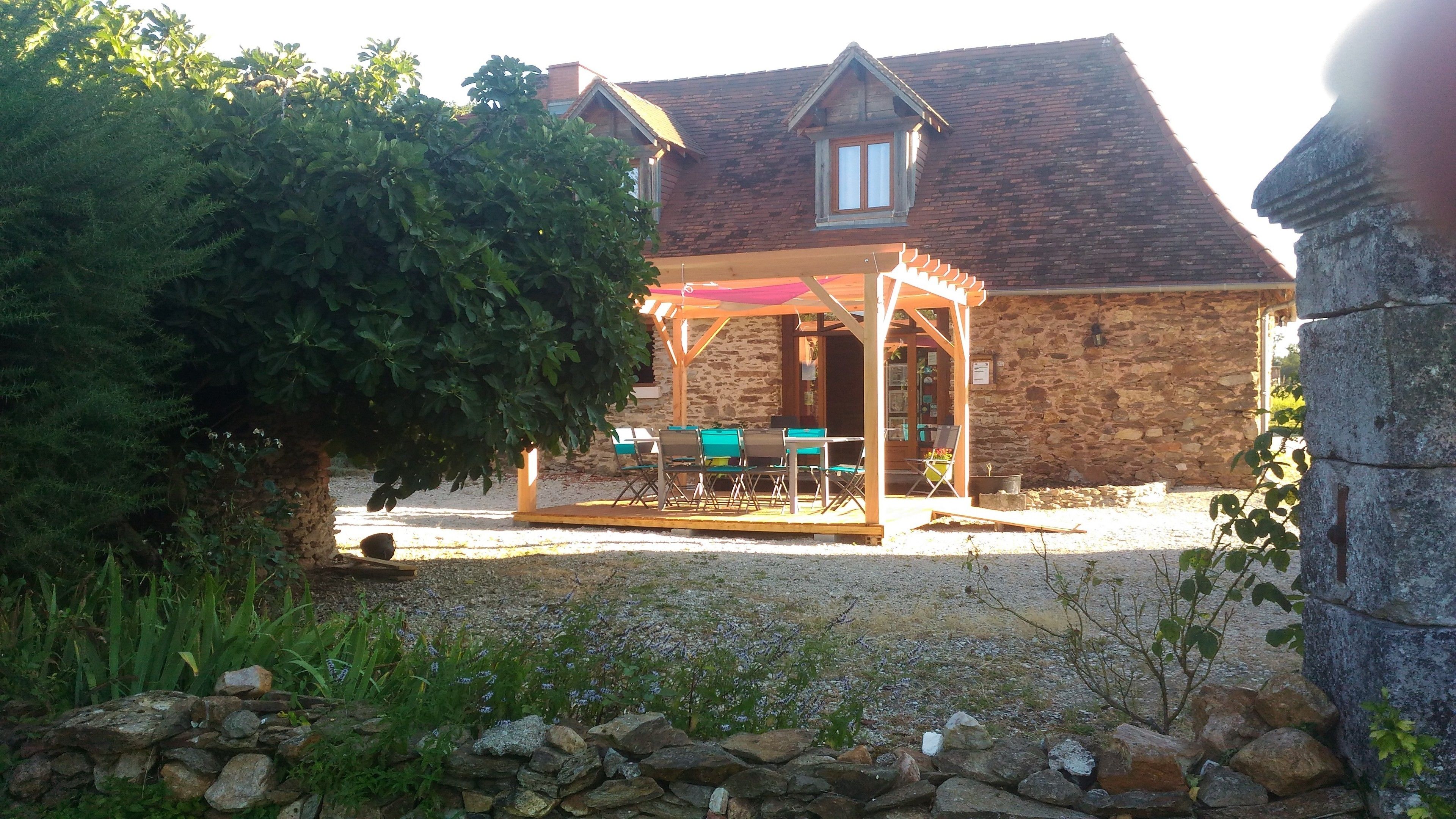 Un edificio de piedra con un porche de madera, una mesa con sillas y un gran árbol en primer plano.