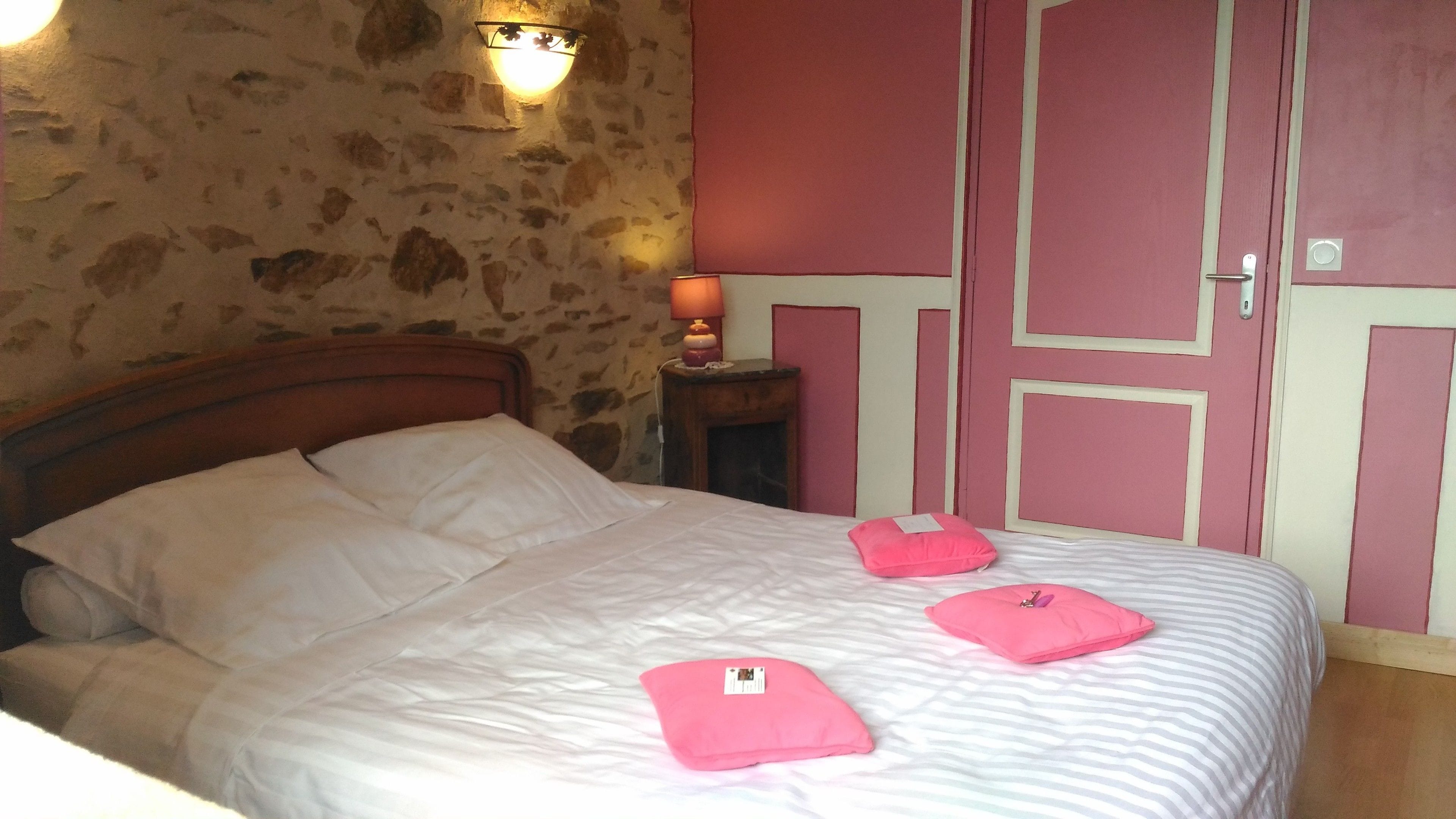 A bed with white linens, pink pillows, and a striped blanket. There is a stone wall and a wooden headboard. A small table with a lamp and a pink pillow is next to the bed.