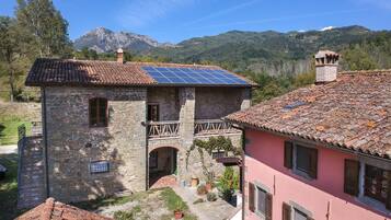 A stone building with a red-tiled roof and a balcony, surrounded by greenery and mountains in the background.