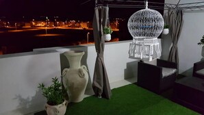 A balcony with a view of a cityscape at night, featuring a large vase, a hanging spherical light fixture, and a small potted plant. - Maryouma Appart (Nabeul)