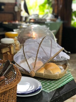 A basket of bread on a checkered cloth, a jar of jam, and a plate with a spoon on a table. - Lua Pousada (Barra de São Miguel)