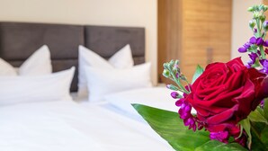 Una cama bien hecha con ropa blanca y almohadas, un cabecero de madera y un ramo de flores rojas y moradas sobre la mesita de noche.