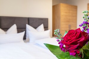Una cama bien hecha con ropa blanca y almohadas, un cabecero de madera y un ramo de flores rojas y moradas sobre la mesita de noche.
