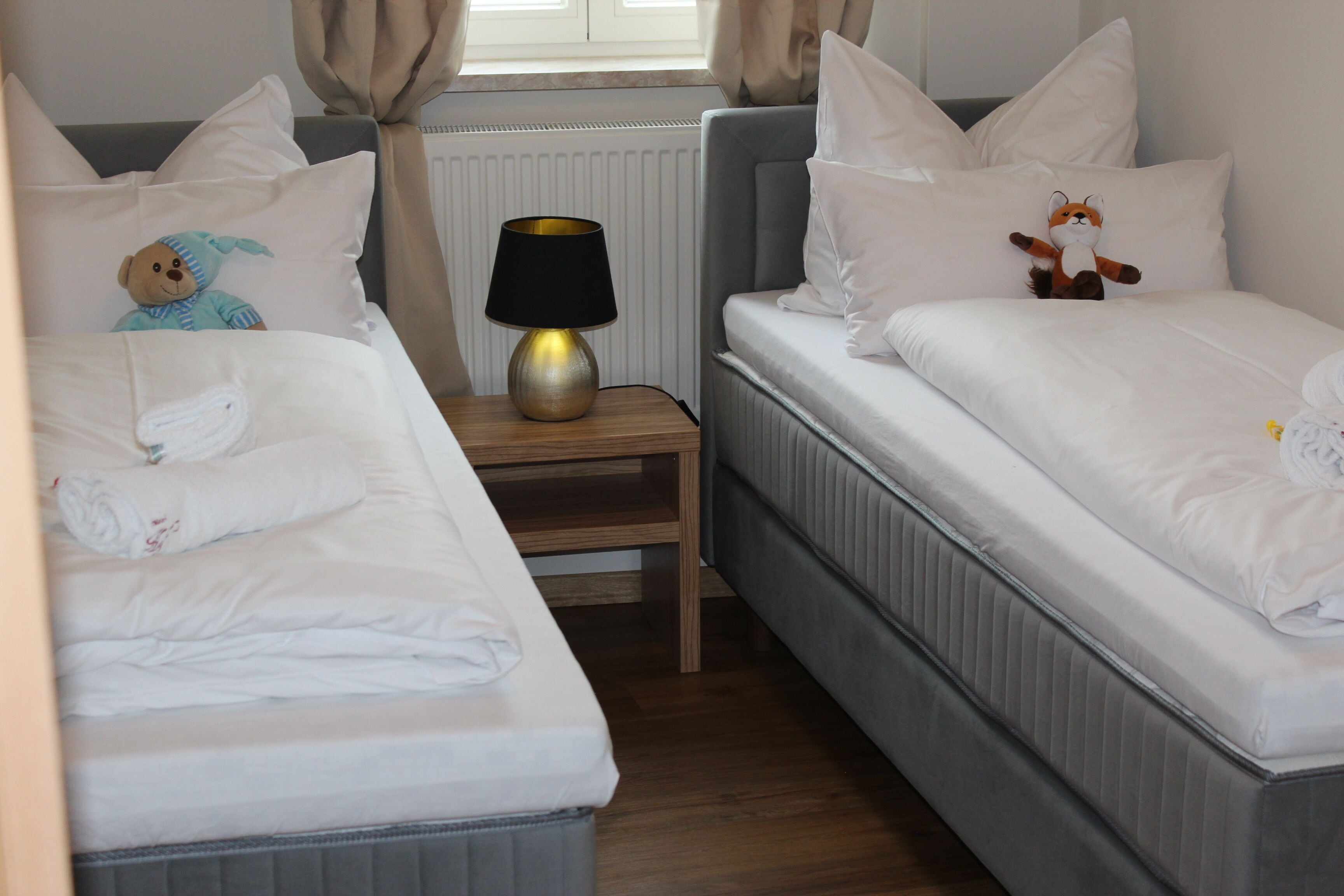 A hotel room with two beds, a bedside table with a lamp, and a window with curtains.