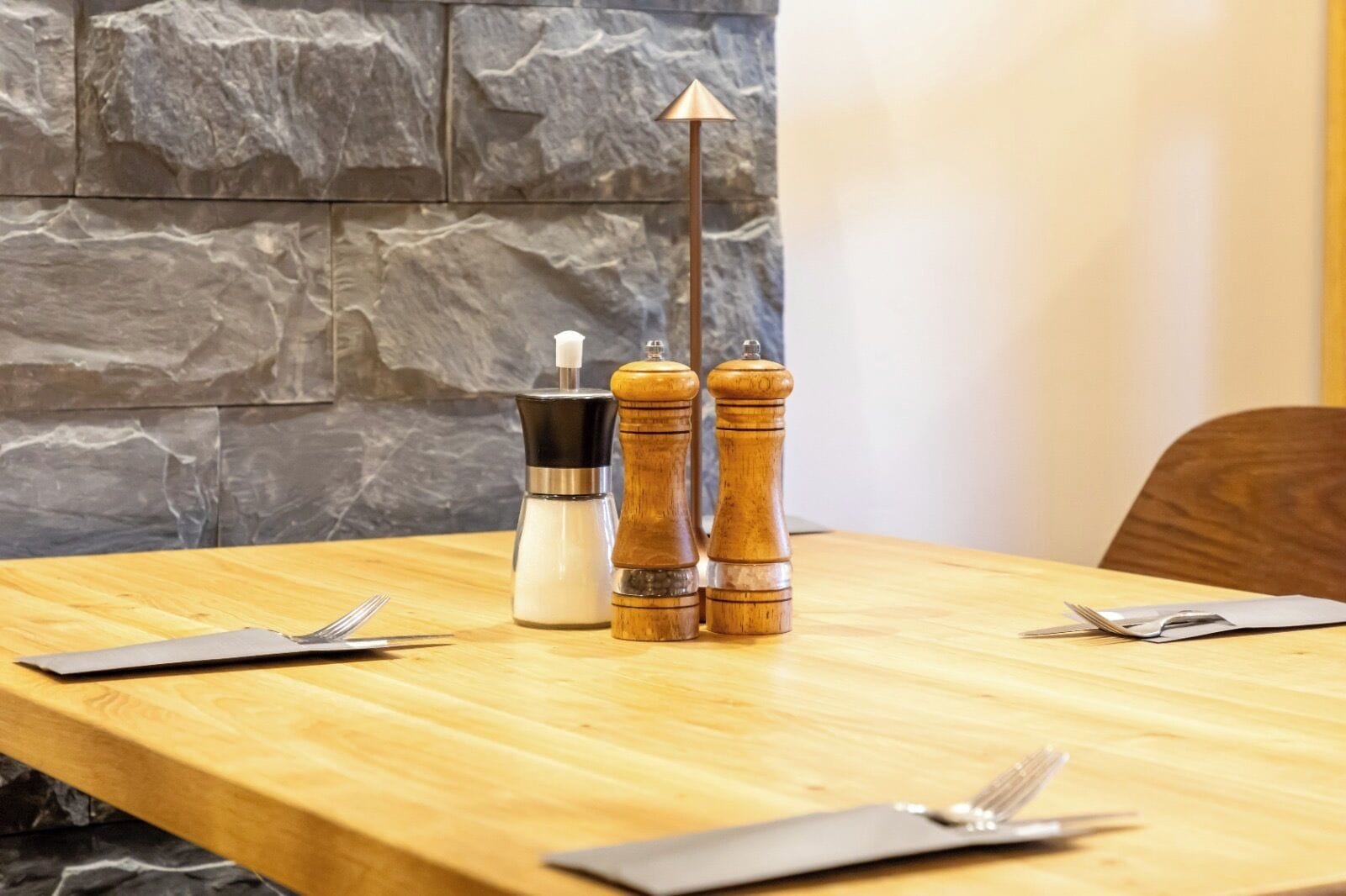 Une table en bois, sur laquelle on trouve un ensemble salière et poivrière, un distributeur de condiments noir et argenté, ainsi qu’un couteau et une fourchette posés sur une serviette.