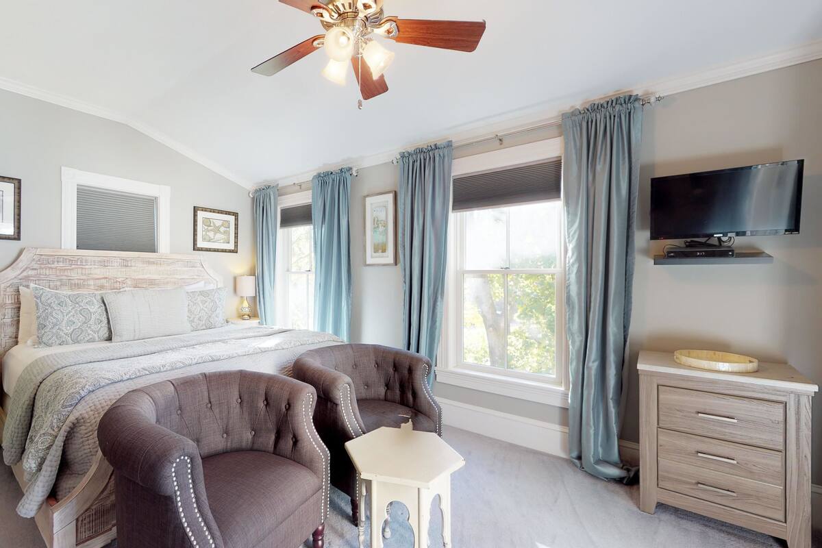 Une chambre à coucher avec un grand lit, deux fauteuils, une petite table, un ventilateur de plafond, une télévision et une fenêtre avec des rideaux.