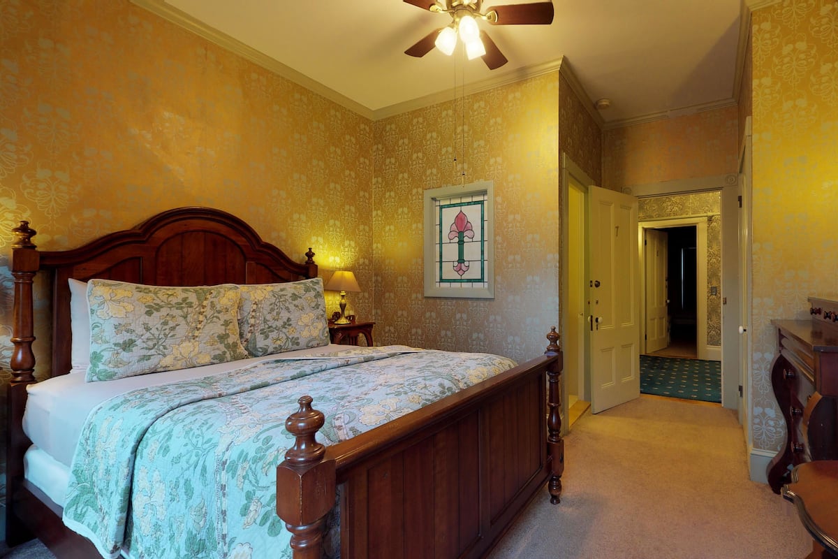 Une chambre à coucher avec un lit en bois, une table de chevet avec une lampe, un miroir et une porte donnant sur une autre pièce.