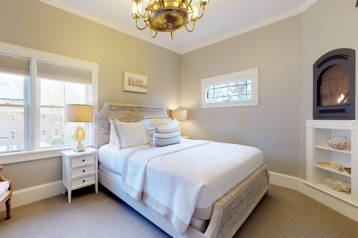 Une chambre avec un grand lit, deux tables de chevet avec des lampes, un fauteuil, une cheminée et une vue sur l’extérieur par les fenêtres.