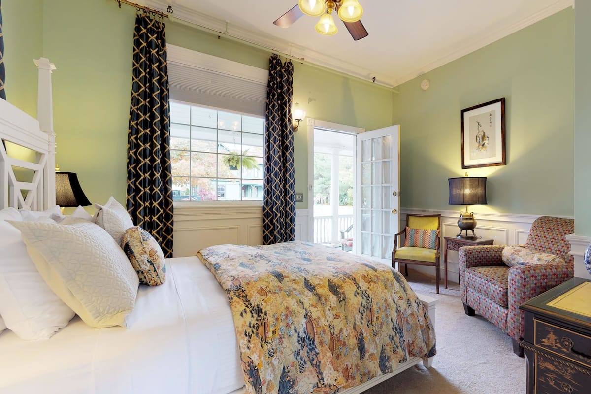 Une chambre à coucher avec un lit, une chaise, une table avec un vase et une fenêtre avec des rideaux.