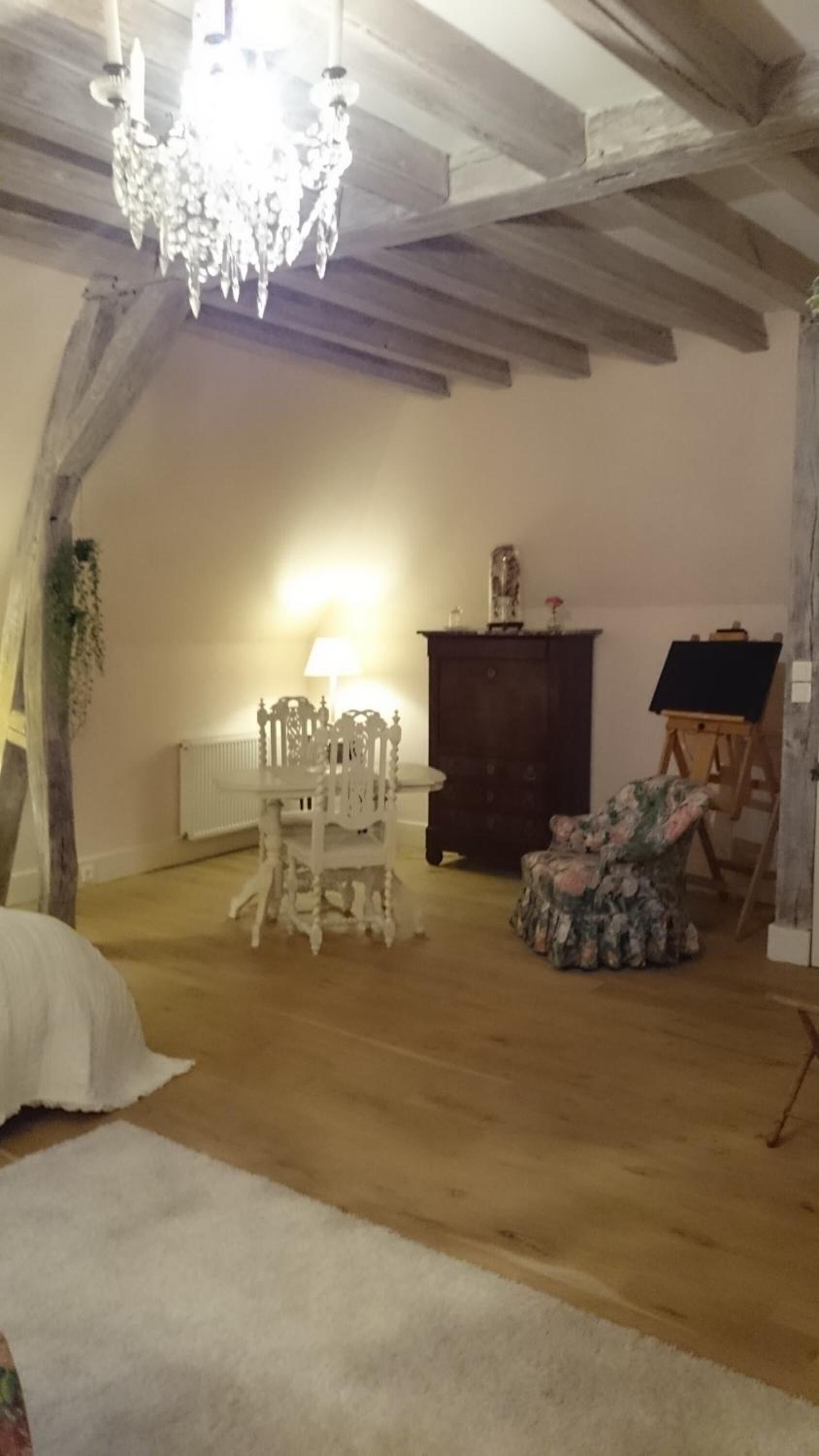 A room with exposed wooden beams, a chandelier, a white chair, a wooden cabinet, and an easel with a blank canvas.