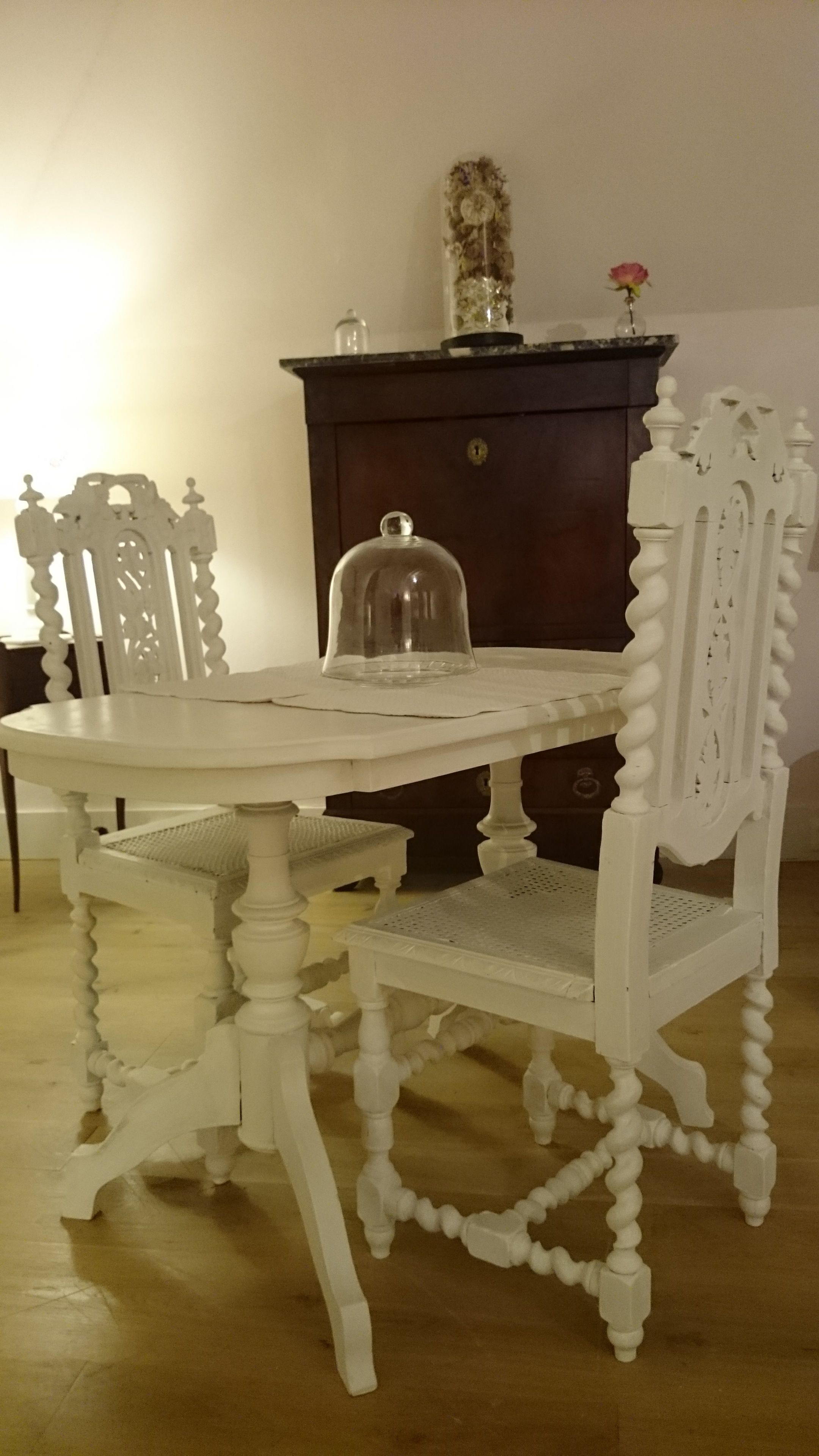 Une table à manger blanche, recouverte d’un couvercle en forme de dôme de verre, est entourée de chaises blanches richement décorées. Un buffet en bois foncé, surmonté d’un élément décoratif, se trouve à l’arrière-plan.