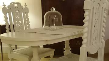 Une table à manger blanche, recouverte d’un couvercle en forme de dôme de verre, est entourée de chaises blanches richement décorées. Un buffet en bois foncé, surmonté d’un élément décoratif, se trouve à l’arrière-plan.