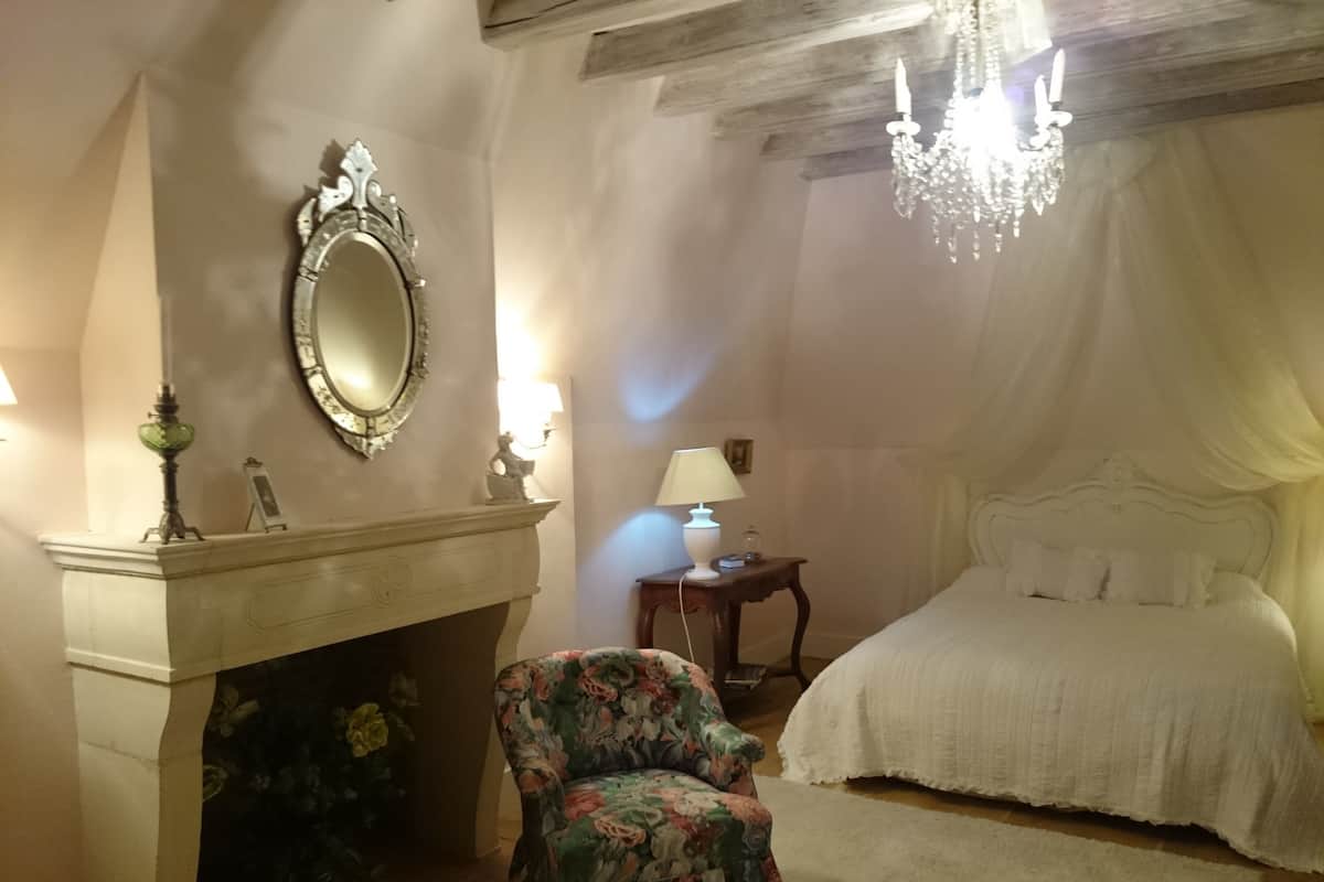 A bedroom with a bed, a chair, a mirror, a fireplace, and a chandelier.