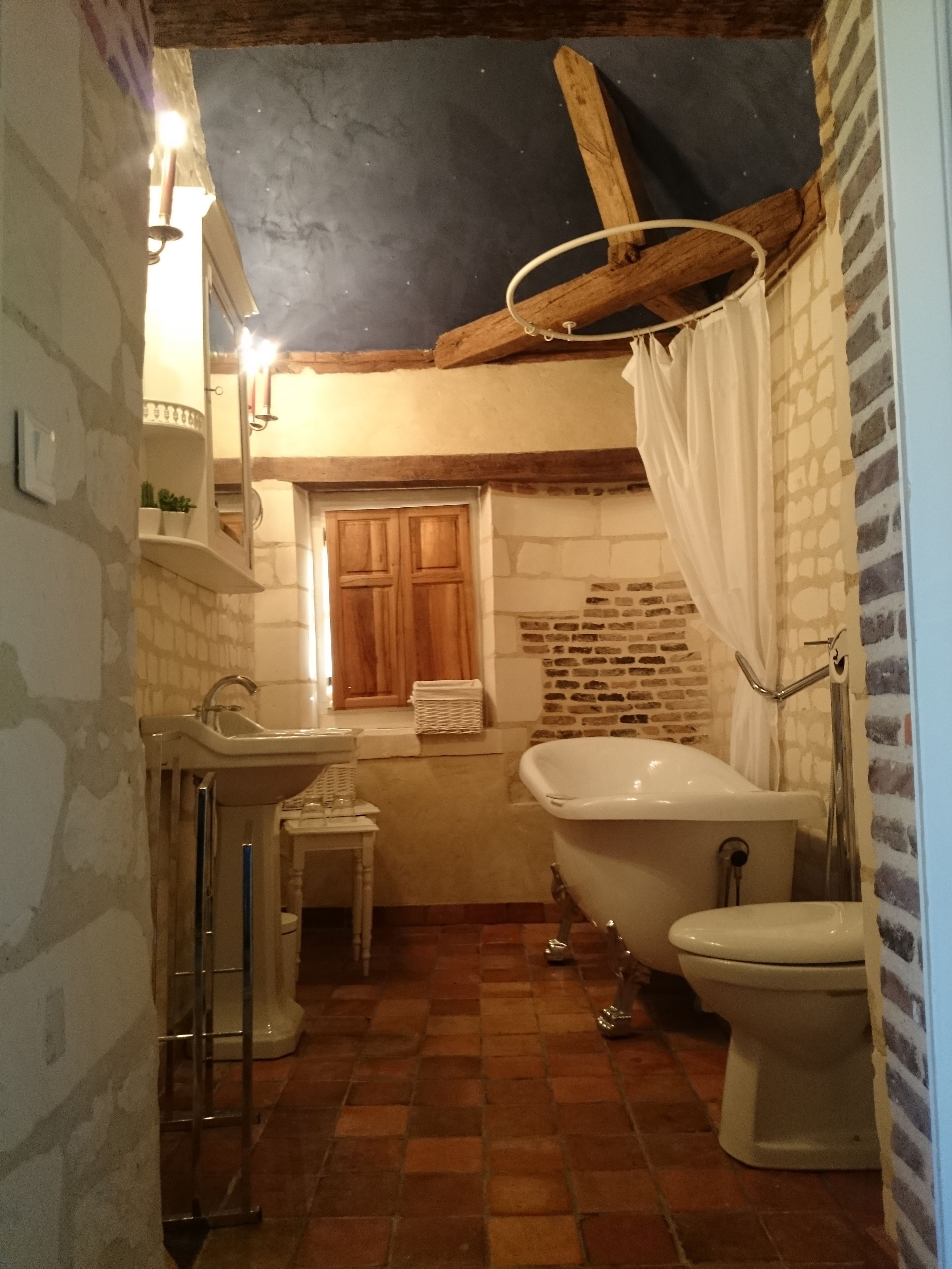 Un baño con bañera, inodoro y lavabo. Tiene una puerta de madera y una ventana con cortina. El piso está cubierto de baldosas.