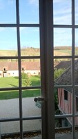 Vista a través de una ventana con celosía metálica, mostrando un paisaje rural con campos, un edificio de techo rojo y cielo despejado.