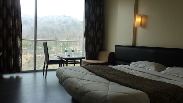 Habitación de hotel con cama, una silla, una mesa con botellas de agua y vistas a un paisaje montañoso a través de un ventanal grande.