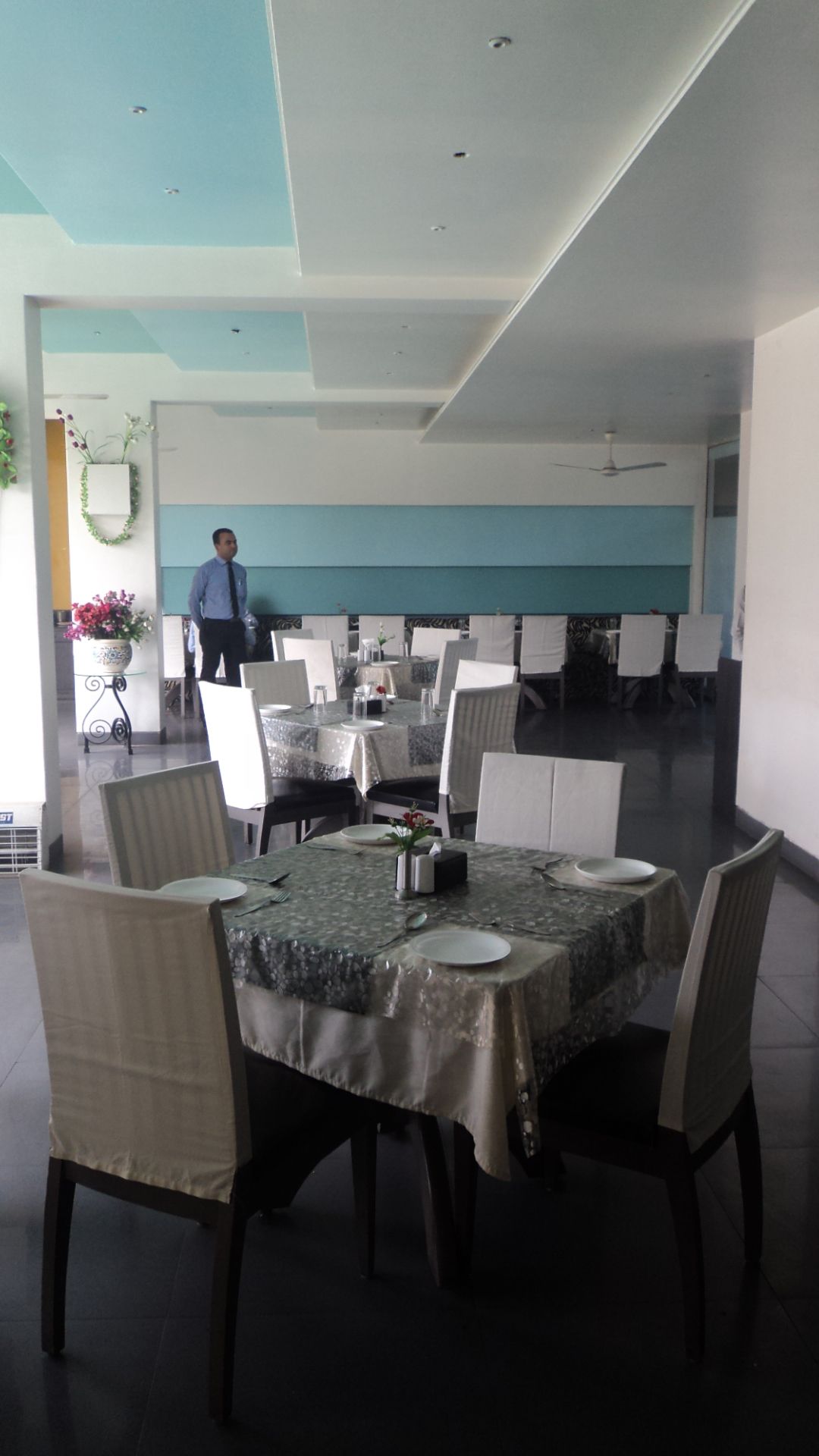A dining area with tables set for four, chairs, a ceiling fan, and a person standing in the background.